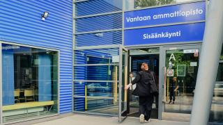 Henkilö kävelee sisään Vantaan ammattiopiston pääsisäänkäynnistä, jonka yläpuolella on kyltti 'Vantaan ammattiopisto Sisäänkäynti'. Rakennuksen julkisivu on sinistä aaltopeltiä ja suuria lasi-ikkunoita.