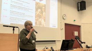 Pirjo Vartiainen avaamassa seminaaria. Taustalla valkokangas, jossa seminaarin ohjelma.